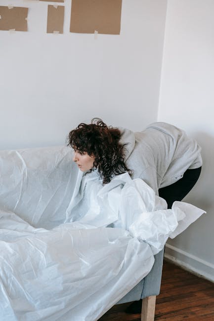 A woman with curly dark hair, dressed in a light grey hoodie and black trousers, is leaning over a white sofa covered with protective white sheets or dust covers. She appears to be in the process of r