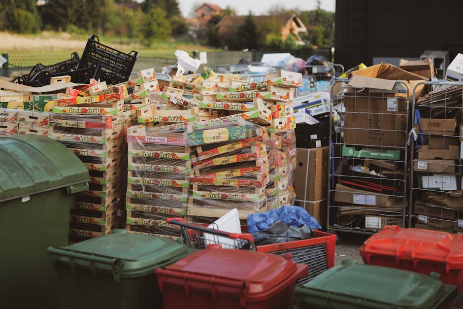 A large green wheeled refuse collection bin is positioned on a paved surface against a dark brick wall. The bin is filled with several red and purple bags of garden compost or similar material, all ma