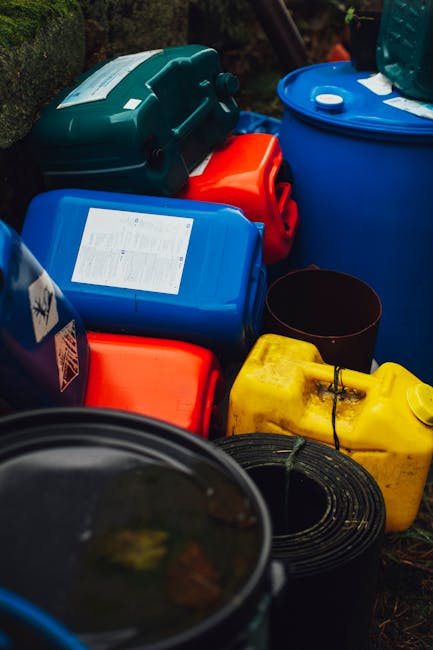 The image shows the tops of four large industrial drum barrels arranged outdoors on a paved pavement section with a patch of grass and small plants visible nearby. The drums are cylindrical with round